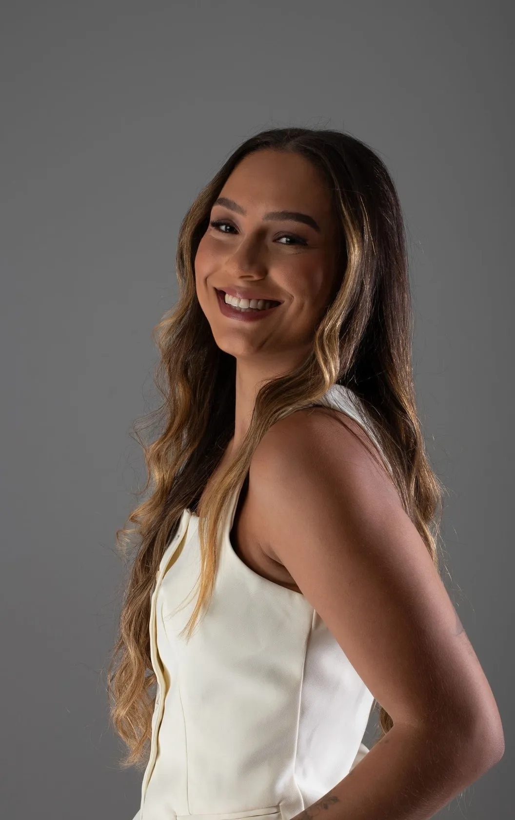 Mulher de cabelos longos sorrindo de perfil em estúdio de fundo cinza.