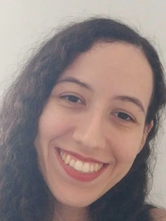 Mylena, de cabelos longos cacheados, sorrindo em frente a uma parede clara.