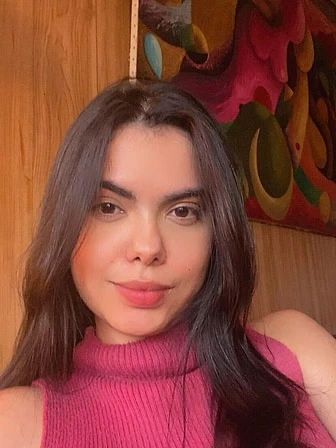 Paula, de cabelos longos escuros e blusa rosa, sorrindo diante de uma parede de madeira com um quadro colorido.